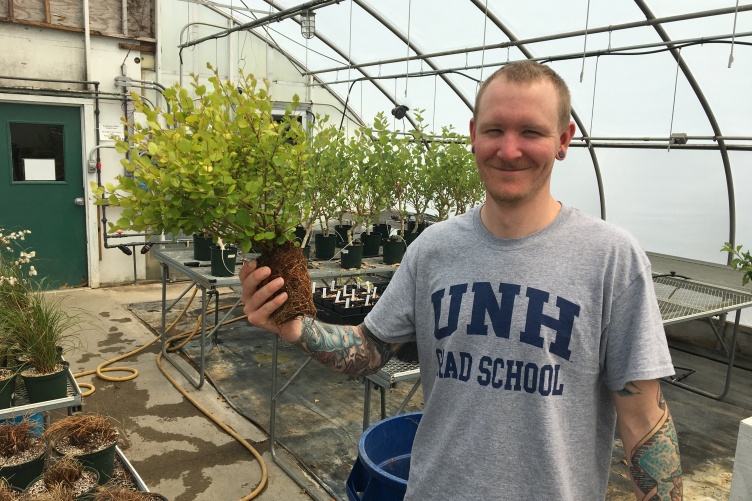 Male student in greenhouse holds a plant. His t-shirt says UNH Graduate School.