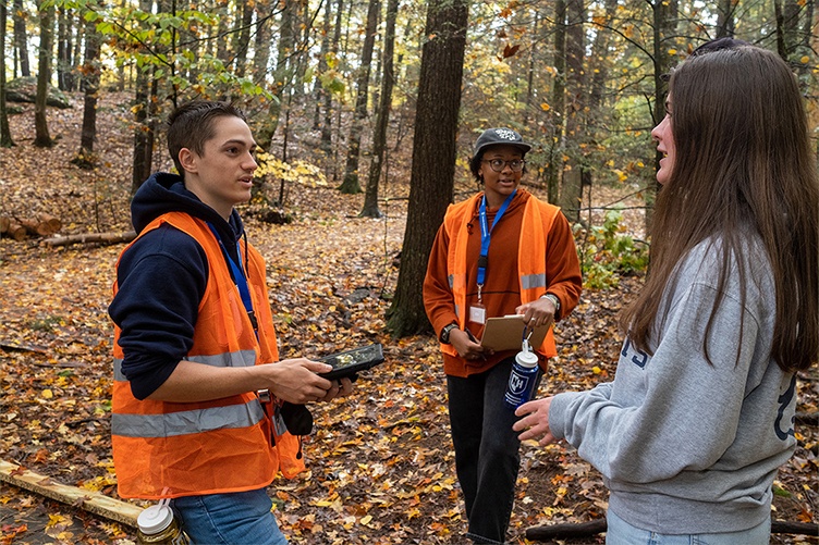 A photo of three students, 2 wearing orange vests, practicing their surveying skills in College Woods.