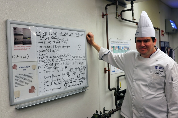 A photo of a UNH Chef Todd Sweet wearing an chef’s outfit stands next to a whiteboard at Holloway Commons with information for staff on what returned items can be composted.
