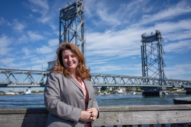Woman with brown hair wearing gray suit stands in front of Memorial Bridge in Portsmouth.