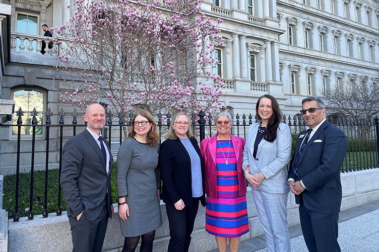 Six people in business attire standing for a photo outside the Eisenhower Executive Office Building in Washington, DC. Behind them is a blooming cherry tree.