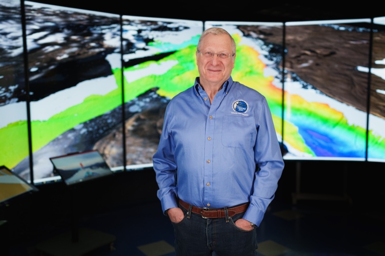 Man in blue shirt stands in front of a screen with mapping graphics on it