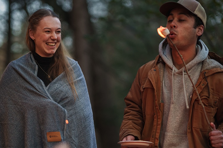 Male student blows out a flaming marshmallow next to a female student wrapped in a blanket