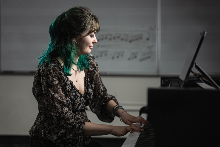 A woman sits at a piano bench and plays, in front of a white board with musical notes on it