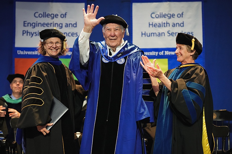 A man waves from the stage as he receives an honorary stole