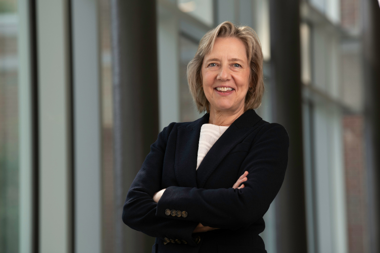 Vanessa Druskat, associate professor of organizational behavior and management at UNH, standing with arms crossed and smiling in a well-lit hallway with large windows.