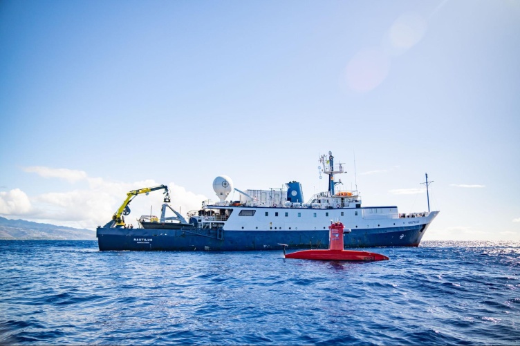 On the open ocean, a small red uncrewed surface vessel floats in front of a much larger research vessel