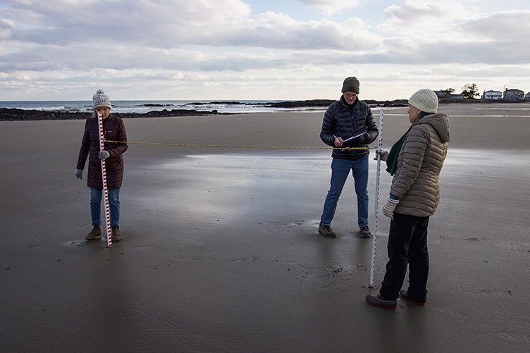 Two women holding yard sticks stand on wet sand in front of the ocean