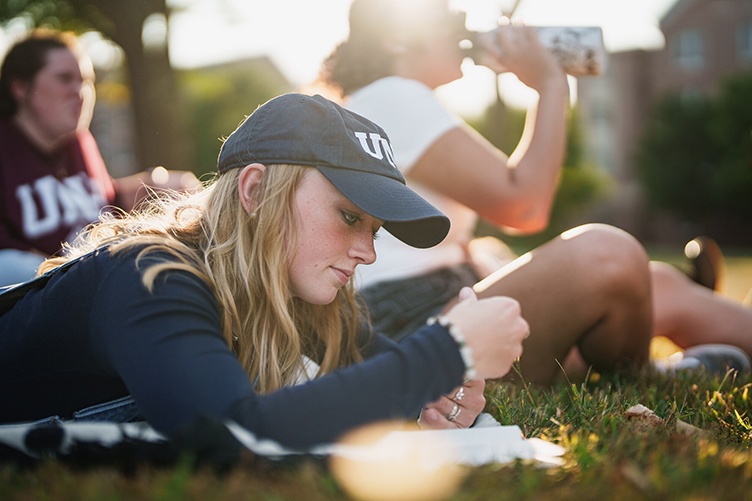Female student lying on her stomach in the grass with two other students in the background