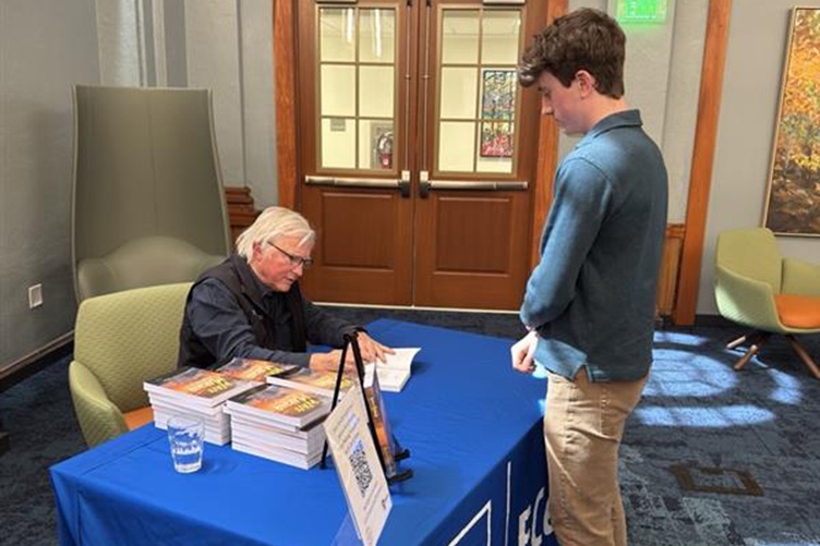 Male student stands in front of a table as a man sits behind it and signs a copy of his book