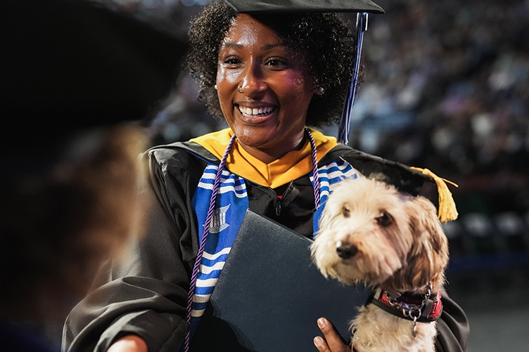 A female graduate holds a white dog in a small grad cap while shaking hands to receiver her diploma