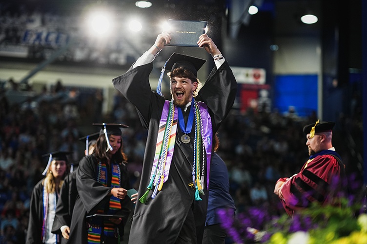 A male graduate hoists his diploma over his head in celebration while walking across the stage