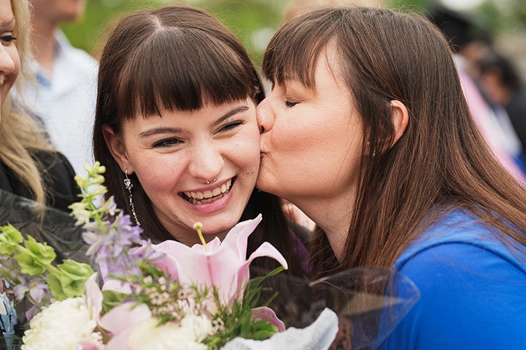 A graduate holding flowers smiles while getting a kiss on the cheek from another woman