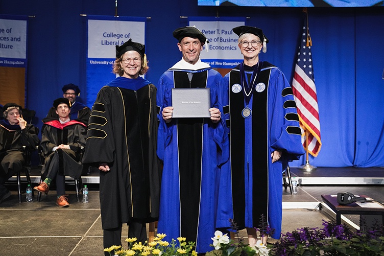 A man holding an honorary diploma stands between two women on stage at commencement