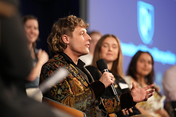 A male student speaks into the microphone during a panel discussion on a stage