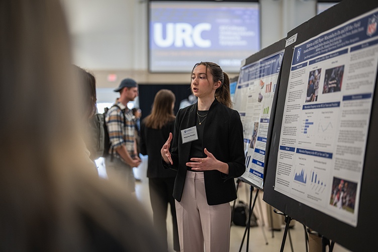 A female student stands in front of a poster explaining its contents to a few people standing in front of her