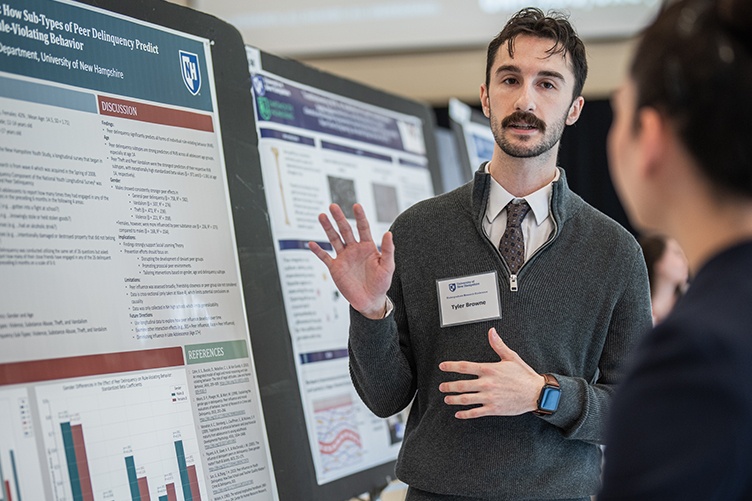 A man with a mustache wearing a sweater over a shirt and tie gestures toward a poster