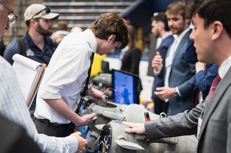 A student in a suit and tie places a hand on a small metal object while speaking to a visitor