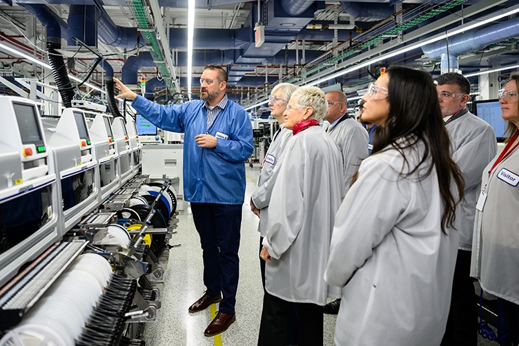 A man in a blue pullover speaks to a small group of people touring a manufacturing floor