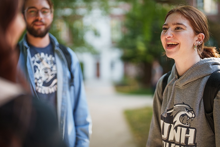 A female student in a gray UNH sweatshirt laughs while talking to two fellow students