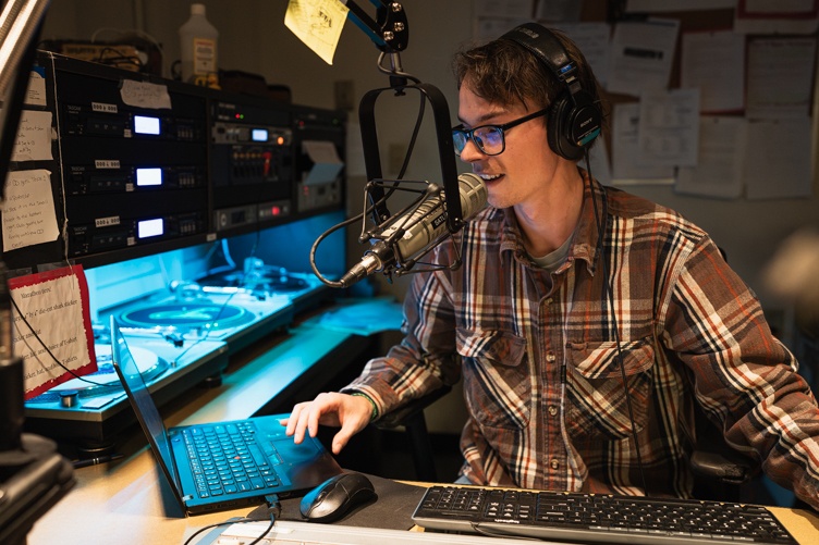 A male student wearing glasses mans the DJ booth in the WUNH studio