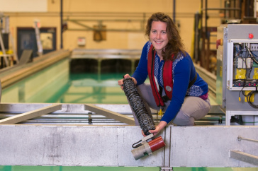 Woman with long brown hair, blue shirt and red life vest stands next to a tank of water while holding a long black cylinder.