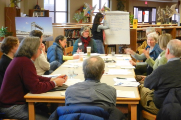 A trained facilitator writes on a large piece of paper while another facilitator leads a discussion at a table of nine participants.