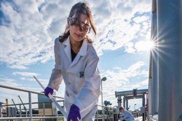 Woman in white lab coat samples liquid outdoors with bright sun over her shoulder