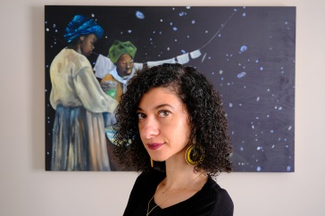Woman with dark curly hair looks at the camera, painting of Black astronomers behind her
