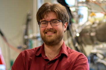 UNH PhD candidate David Ruth smiles at the camera in front of physics equipment