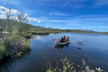 Four people sit in a row boat on a lake surrounded by green vegetation.