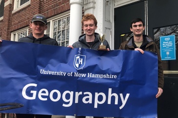 three students holding trophy and geography banner