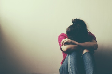 A young woman sits against a blank wall with her head on her knees.