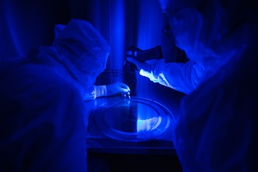 Two people in white Tyvek suits examine a thin flat disk under a blue ultraviolet light that detects dust.