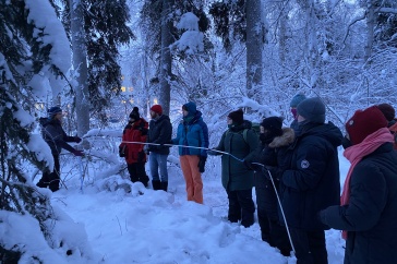 People bundled up in winter gear hold a long white tube to measure soil frost amidst a snowy forest. 