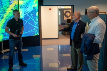 Three men stand in front of a colorful data visualization screen