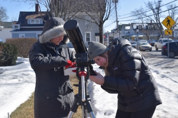 Two female students dressed in warm winter gear try to set up a telescope outside near a snow-covered sidewalk.