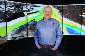 Man in blue shirt stands in front of a screen with mapping graphics on it