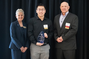 A man holding an award stands between a woman on the left and a man on the right