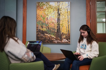 Two women students sit across from each other in chairs, working on laptops