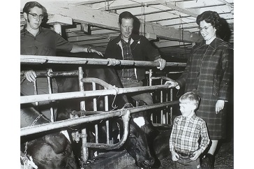 Two men and a woman stand with a child in a dairy barn in a black and white old photo