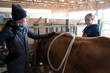 Two woman stand next to the rear side of a cow. Both are smiling. One woman holds a tubing around the cows back.