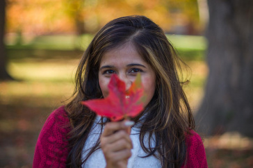 A UNH student holding a red leaf from a maple tree, with golden hues of foliage in the background