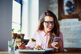 a woman looking at her cell phone while eating BARANQ/SHUTTERSTOCK