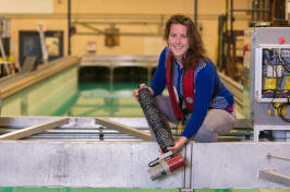 Woman with long brown hair, blue shirt and red life vest stands next to a tank of water while holding a long black cylinder.