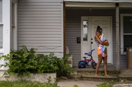 A woman standing in front of her house, holding a child wrapped in a towel.