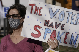 A woman in a mask and glasses hold a sign that says "the voters have spoken"