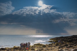 Sun peeks through clouds and a group of students stands on a seaweed-covered rocky coastline by the ocean.