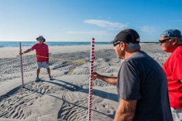 Three elderly men stand on a beach holding measuring sticks to determine height of the sand.