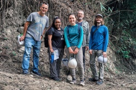 A photo showing five people standing in front of a cliff face. The people are mix of men and women and several of them wear mining helmets.